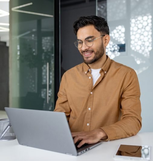 Indian young man studying and working in a modern office, sitting at a desk, smiling and typing on